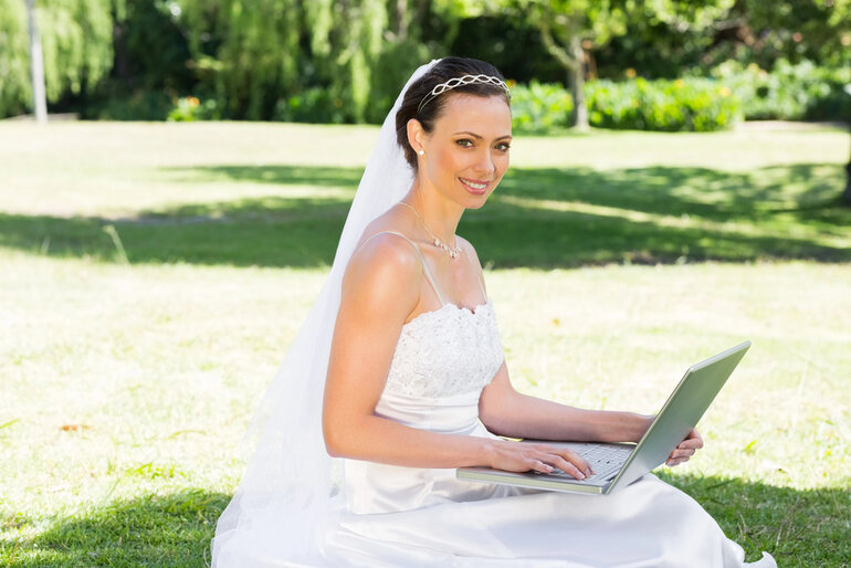 Smiling bride in a wedding dress using a laptop outdoors, symbolizing online matchmaking and international marriage topics.